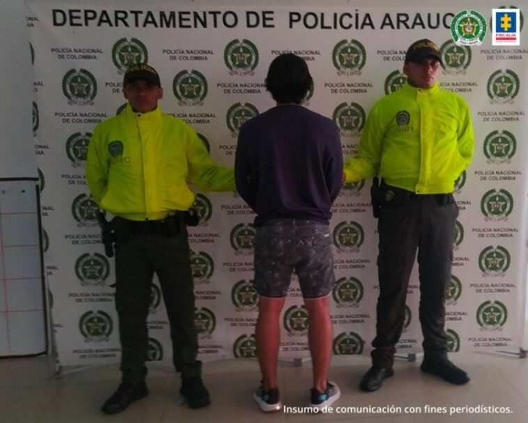 El capturado aparece de espalda con ropas oscuras. Dos personas de la Policía Nacional lo vigilan a cada lado. Al fondo hay un pendón de la Policía.