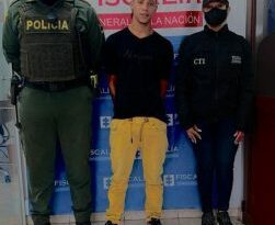 Jaider Suárez Bedoya aparece en la foto con los brazos a la espalda;  vestido con una camiseta negra, pantalones mostaza y zapatillas blancas y negras.  A su derecha un oficial de la Policía Nacional uniformado ya su izquierda un servidor del CTI del Ministerio Público.  Detrás de ellos aparece una pancarta institucional de la Fiscalía.