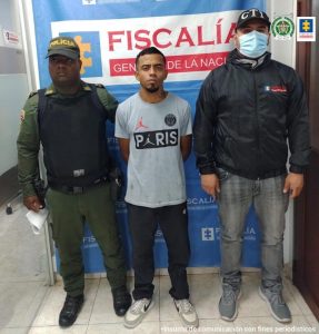 En la foto aparece Sebastián Vélez Cardona, con los brazos atrás; vestido con camiseta gris y letras estampadas, pantalón oscuro y tenis. Al lado derecho aparece un uniformado de la Policía Nacional y al lado izquierdo un servidor del CTI de la Fiscalía. Detrás de ellos hay un pendón institucional de la Fiscalía.