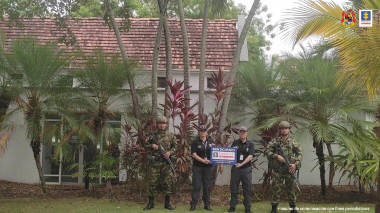 Al fondo es´ta la vivienda afectada con las medidas cautelares. En frente hay dos personas del CTI sosteniendo un cartel que dice Fiscalía y a cada lado hay una persona del ejército.