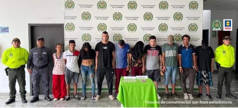 En la foto aparecen de pie y en fila los presuntos integrantes del grupo delictivo ‘Los de Papá’. Al lado derecho de ellos hay un uniformado de la Policía Nacional y un servidor del CTI de la Fiscalía. A la izquierda de ellos hay un uniformado de la Policía. Delante de ellos hay una mesa con mantel verde con varias bolsas plásticas pequeñas y dinero en efectivo, así como dos letreros que dicen ‘bazuco’ y ‘dinero’. Detrás de ellos hay un pendón institucional de la Policía.