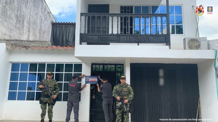 Al fondo, la casa afectada por las medidas cautelares.  Al frente hay dos personas del CTI con un cartel que dice Fiscalía, ya los lados hay una persona del ejército.