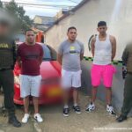 En la imagen se observan tres hombres en bermudas, tenis y camisetas custodiados por dos agentes de la Policía Nacional delante de un carro que fue in inmovilizado.