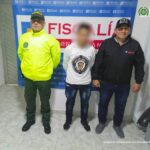 En las fotografía aparece un capturado junto a personal de Fiscalía General de La Nación y Policía Nacional. En la parte posterior de la imagen se ven banners de Policía Nacional y la Fiscalía General de la Nación.