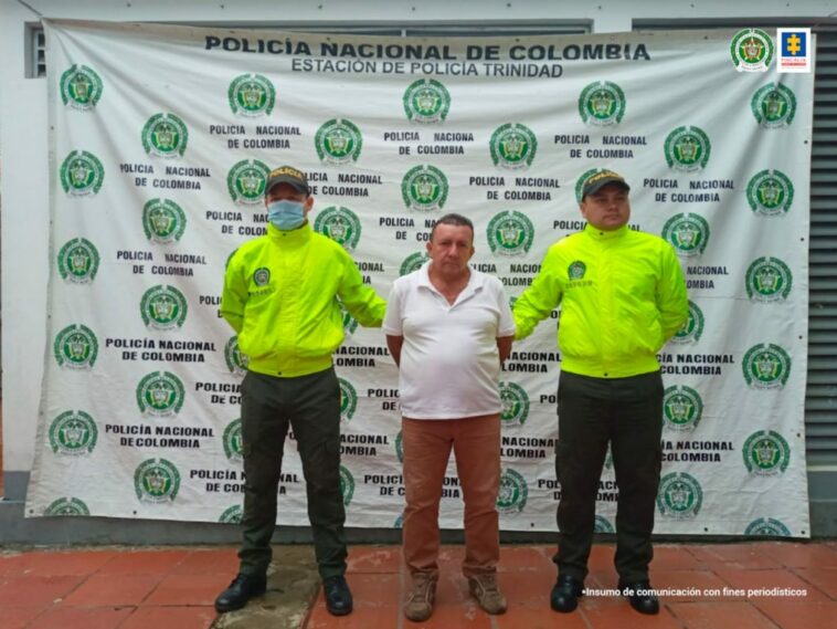 El capturado esta custodiado por dos uniformadios de la Policía Nacional, está con sus manos esposadas a la espalda y de fondo un pendon institucional.