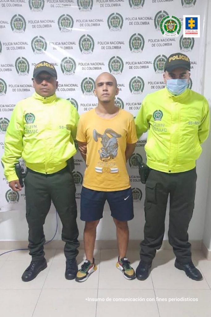 En la imagen se observa a un hombre de camiseta amarilla y bermuda oscura custodiado por dos agentes de la Policía Nacional delante de un pendón de esa institución.