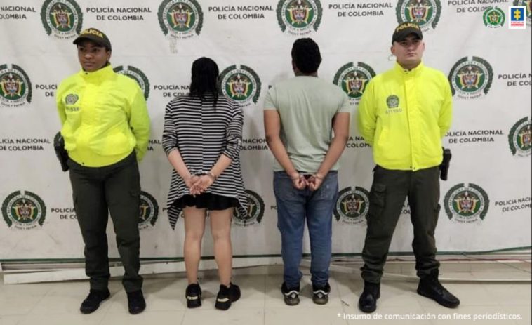 En la imagen se   a una pareja de espalda custodiada por dos agentes de la Policía Nacional delante de un pendón de esa institución.
