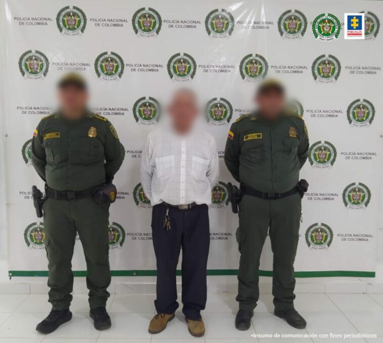 En la imagen se observa a un hombre con camisa blanca y pantalón oscuro custodiado por dos uniformados de la Policía Nacional, delante de un pendón de esta institución.