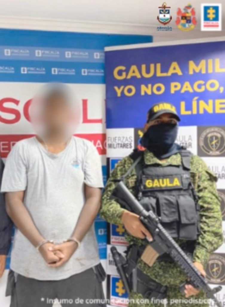 En la imagen se observa a un hombre esposado en sus manos con camiseta blanca y bermuda gris. A su lado se encuentra un uniformado del Ejército Nacional delante de dos pendones, uno de la Fiscalía y el otro del Ejército Nacional.