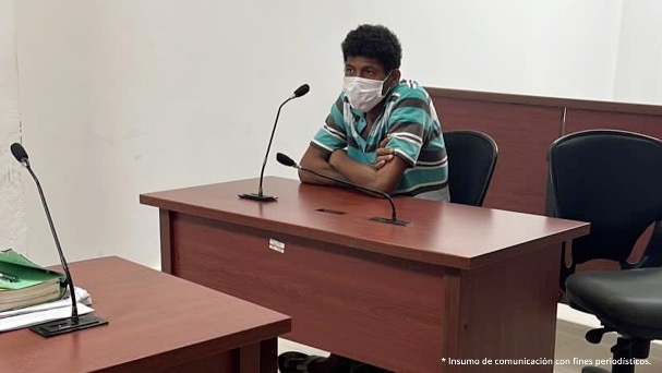 En la fotografía se observa a un hombre sentando detrás de un escritorio, con tapabocas, camiseta azul y rallas cafés, tiene los brazos cruzados y con un micrófono en la boca.