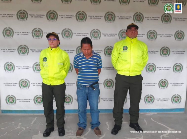 Se observa un hombre vestido con camiseta de rayas horizontales en azul y blanco, de frente y a cada costado un funcionario de Policía Nacional.  Al fondo un pendón de Policía Nacional