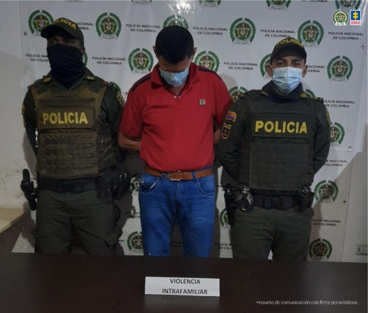 En la foto aparece Jesús Antonio Ríos Castrillón, con tapabocas, los brazos atrás y la cabeza abajo. Vista camiseta roja y jean azul. A lado y lado de él hay dos uniformados de la Policía Nacional. Delante de ellos hay una mesa con mantel verde y sobre ella un letrero que dice: ‘Violencia intrafamiliar’. Detrás de ellos hay un pendón institucional de la Policía.