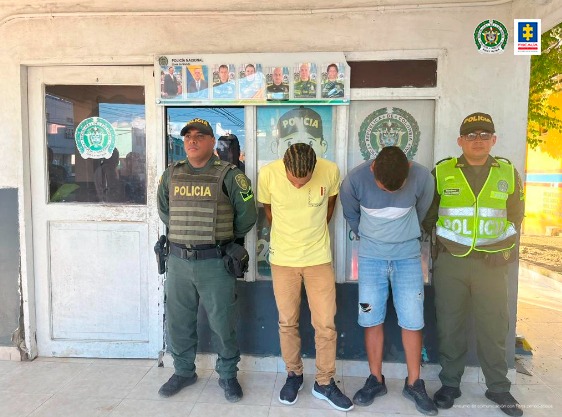 En la imagen se observan a dos hombres uno de camiseta amarilla y pantalón beis y el otro de camiseta azul con jean custodiados por dos agentes de la Policía Nacional.