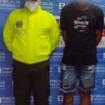 En la imagen se observa a un hombre de camiseta negra, bermuda de jean y chanclas negras, acompañado por un uniformado de la Policía Nacional,  delante de un pendón de la Fiscalía General de la Nación.