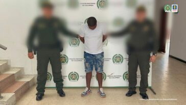 En la imagen se observa a un hombre de camiseta blanca con bermuda de jean y tenis, custodiado por dos agentes de la Policía Nacional delante de un pendón de esa institución.