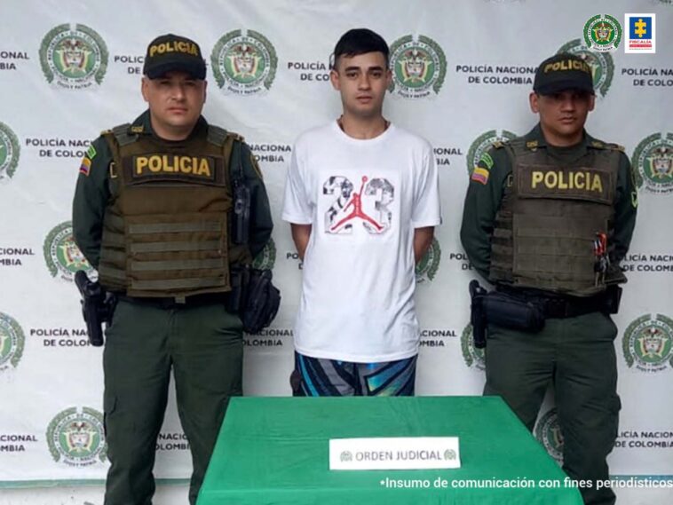 Se observa al centro el capturado y a cada uno de sus costados un funcionario de Policía Nacional.  Al fondo un pendón de Policía Nacional.  Al frente una mesa con el cartel 'ORDEN JUDICIAL.