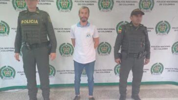 En la foto aparece, de pie y con  los brazos atrás, Cristian Augusto Colorado Caro. Vista camiseta blanca de manga corta, jean azul y zapatos oscuros. A lado y lado de él hay uniformados de la Policía Nacional.  Detrás de ellos hay un pendón institucional de la Policía.