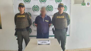En la imagen se observa a un hombre vestido de camiseta azul y jean custodiado por dos agentes de la Policía Nacional, delante de un pendón de esa institución.