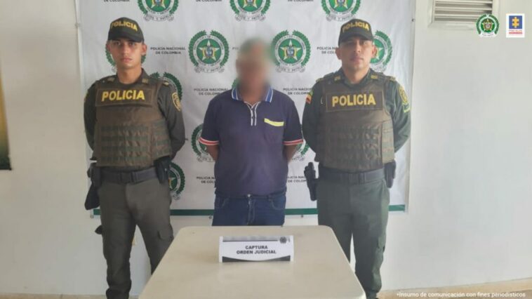 En la imagen se observa a un hombre vestido de camiseta azul y jean custodiado por dos agentes de la Policía Nacional, delante de un pendón de esa institución.