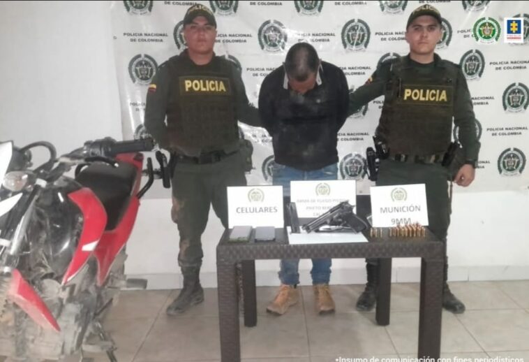 En la fotografía aparece el capturado junto a dos agentes de la Policía Nacional. En la parte posterior se observa una mesa con un arma de fuego, munición y dos teléfonos celulares.