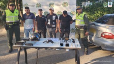 En la imagen se observa a cuatro hombres custodiados por dos agentes de la Policía Nacional, detrás de una mesa donde se identifican un arma de fuego, municiones y celulares.