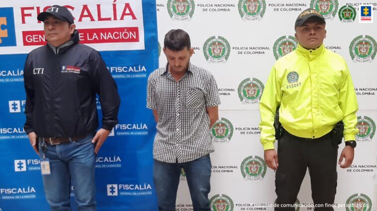 El capturado aparece frente a un banner de la Fiscalía. Tiene a sus lados a una persona de la Fiscalía y de la Policía.