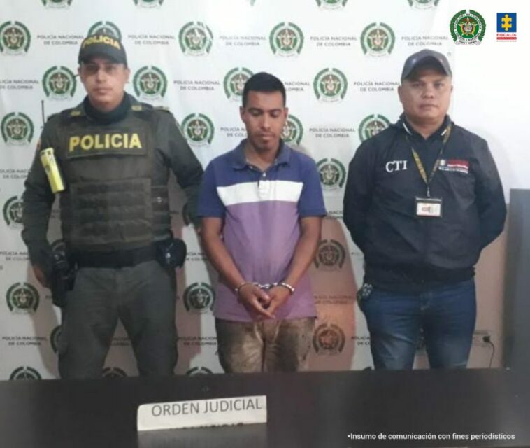 En la foto aparece, de pie, Yulián Andrey Ramírez Carvajal, con los brazos adelante y esposado. Viste camisa color lila con azul, con franja blanca y manga corta; así como pantalón café claro. Al lado derecho de él hay un uniformado de la Policía Nacional. Al lado izquierdo de él hay un servidor del CTI de la Fiscalía. Delante de ellos hay una superficie oscura y sobre ella un letrero que dice ‘orden judicial’. Detrás de ellos hay un pendón institucional de la Policía.