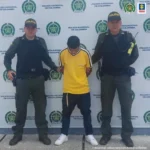 En la foto, José Wilder Zúñiga aparece de pie con los brazos detrás de él.  Viste una camisa amarilla, pantalones oscuros y tenis blancos.  A ambos lados de él hay uniformes de la Policía Nacional.  Detrás de ellos hay una pancarta institucional policial.