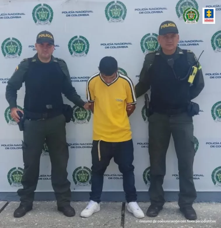 En la foto, José Wilder Zúñiga aparece de pie con los brazos detrás de él.  Viste una camisa amarilla, pantalones oscuros y tenis blancos.  A ambos lados de él hay uniformes de la Policía Nacional.  Detrás de ellos hay una pancarta institucional policial.