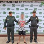 capturado de frente en pantaloneta y chancletas custodiado por dos uniformados de la Policía. Detrás banner de la Policía.