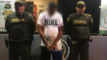 En la fotografía se observa a un hombre con camiseta blanca, bermuda azul, sandalias blancas custodiados por dos agentes de la Policía Nacional en un CAI de Cartagena.