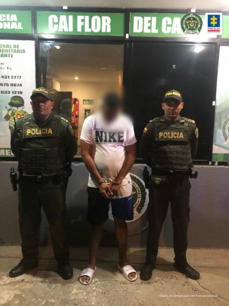 En la fotografía se observa a un hombre con camiseta blanca, bermuda azul, sandalias blancas custodiados por dos agentes de la Policía Nacional en un CAI de Cartagena.