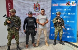 Capturado de frente custodiado por investigador del CTI de la Fiscalía y dos uniformados del Ejército Nacional. Detrás banner de Fiscalía y de Ejército.