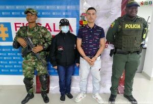 En la fotografía se observa al capturado junto a un servidor del CTI, un agente de la Policía Nacional y un soldado del Ejército Nacional. En la parte superior se observan un banner de la Fiscalía General y otro banner del Ejército Nacional