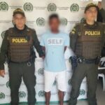 En la fotografía se observa a un hombre moreno, con una camiseta azul clara, bermuda blanca, sandalias negras, custodiado por dos agentes de la Policía Nacional, delante de un pendón de esa institución.