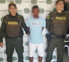 En la fotografía se observa a un hombre moreno, con una camiseta azul clara, bermuda blanca, sandalias negras, custodiado por dos agentes de la Policía Nacional, delante de un pendón de esa institución.