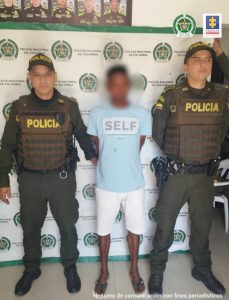 En la fotografía se observa a un hombre moreno, con una camiseta azul clara, bermuda blanca, sandalias negras, custodiado por dos agentes de la Policía Nacional, delante de un pendón de esa institución.