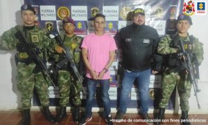 El capturado tiene una camiseta rosada, está esposado y custodiado por dos hombres, uno del CTI de la Fiscalía y el otro un miembro del ejército de Colombia.