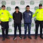 En la fotografía se observan cuatro personas, dos de ellas los capturados y dos funcionarios de la Policía Nacional. En la parte posterior se encuentra un backing con escudos de la Policía Nacional.