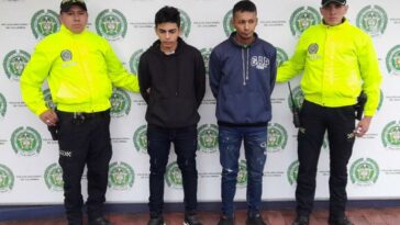 En la fotografía se observan cuatro personas, dos de ellas los capturados y dos funcionarios de la Policía Nacional. En la parte posterior se encuentra un backing con escudos de la Policía Nacional.