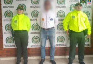En la imagen el investigado esta de frente, esposado con las manos atrás y custodiado por dos uniformados de la Policía Nacional.