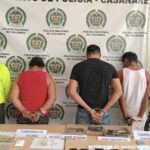 los capturados de espaldas custodiados por dos uniformados de la Policía, delante de ellos una mesa con material incautado, detrás banner de la Policía