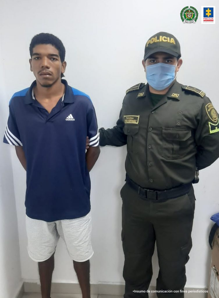 En la fotografía se observa a un hombre moreno con las manos atrás, de camiseta azul oscura con blanca y bermuda blanca, custodiado por un agente de la Policía Nacional.