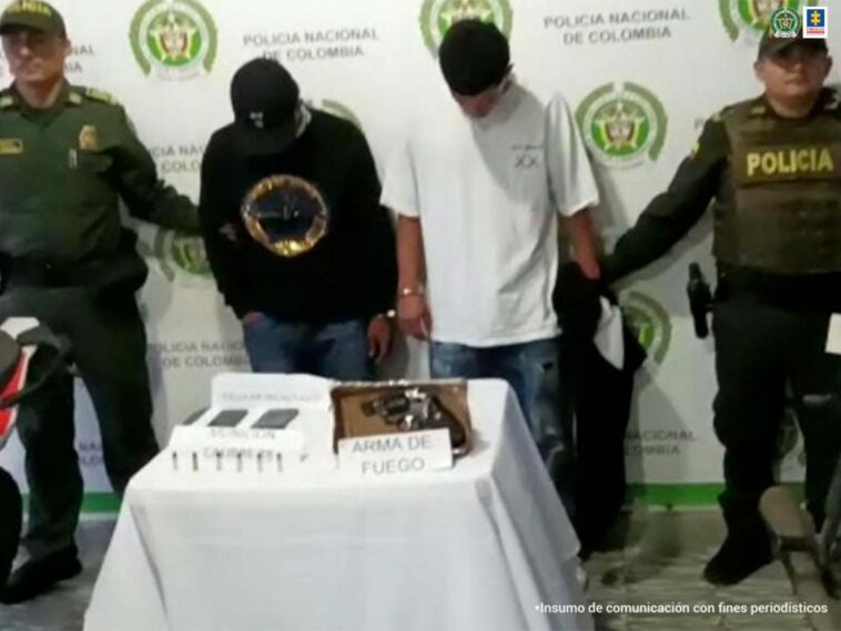 En la foto aparecen de pie y la cabeza agachada, Royer Alberto Naranjo y Hernán Santiago Jiménez Hernández. Uno de los imputados viste buso negro con estampado en el pecho, jean azul y gorra oscura. El otro imputado viste camiseta blanca de manga corta y jean azul. A lado y lado de ellos hay uniformados de la Policía Nacional. Delante de ellos hay una mesa con mantel blanco y sobre ella hay un arma de fuego, dos celulares y munición. Al lado derecho e izquierdo de la mesa hay dos motocicletas. Detrás de ellos hay un pendón institucional de la Policía.