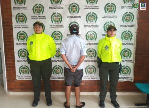 El capturado está esposado de espaldas, custodiado por dos uniformados de la Policía Nacional. Detrás banner de la Policía.