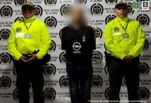 En la foto se ve una persona detenida, custodiada por dos integrantes de la Policía Nacional. Detrás suyo un backing de la institución.