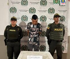En la imagen se observa a un hombre con camiseta negra y pantalón negro con la cabeza agachada, custodiado por dos agentes de la Policía Nacional delante de un pendón de esa institución.