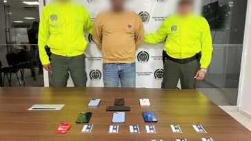 En la imagen se observa a un hombre de buso amarillo y jean azul claro custodiado por dos agentes de la Policía Nacional. Delante de él se identifica una mesa con celulares y tarjetas de celulares.