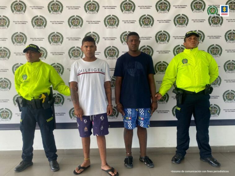 En la imagen aparece un hombre de camiseta blanca con bermuda azul al lado de otro hombre de camiseta azul oscura y bermuda azul con blanca custodiados por dos agentes de la Policía Nacional, delante de un pendón de esa institución.