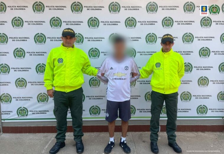 capturado en pantaloneta y camiseta esposado con las manos atrás, custodiado por dos uniformados de la Policía Nacional. Detrás banner de la institución.